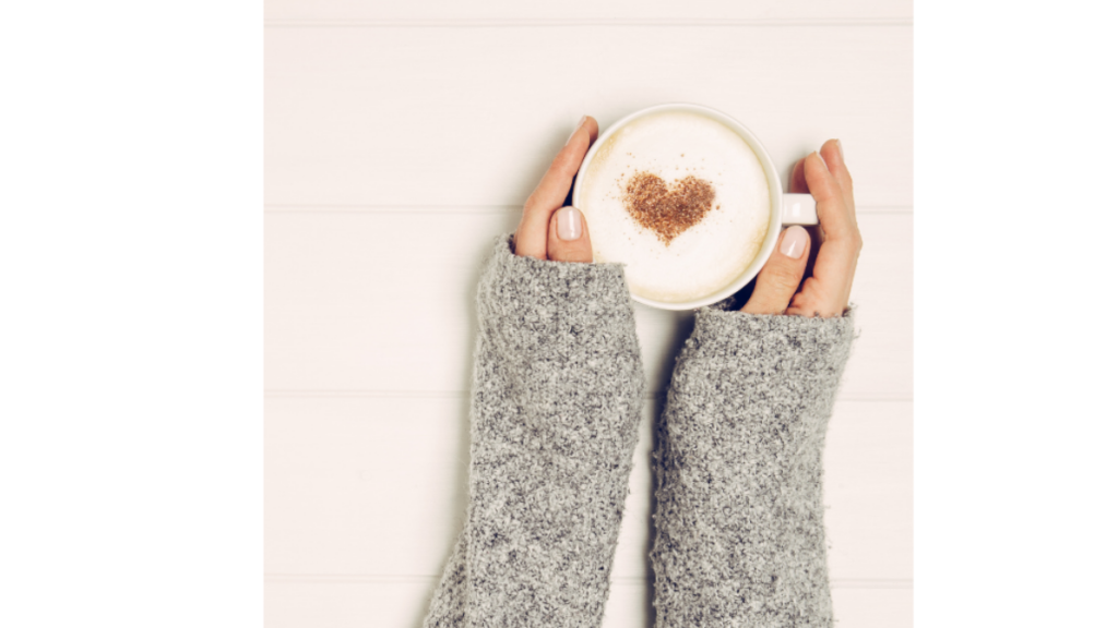 ladies hands in a grey jumper wrapped round a mug with heart shape piped into the drink