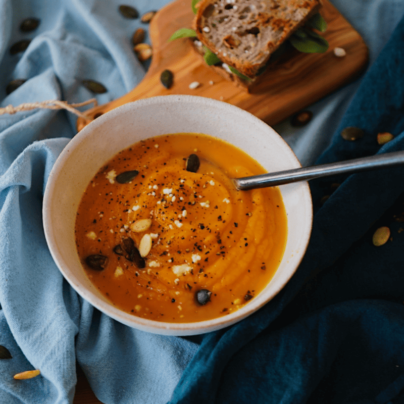 pumpkin season  pumpkin soup in a white bowl on a blue napkin. Granary bread on a rustic bread board in the background 